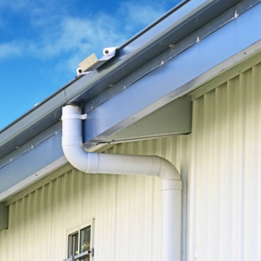 Light-colored commercial metal building in Atlanta, GA with Spartina seamless gutters and a curved downspout along the eaves, set against a bright blue sky.