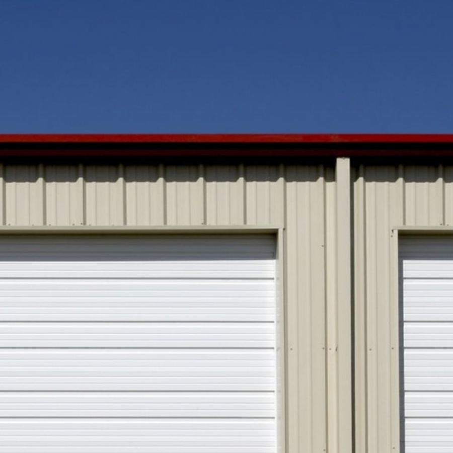 Commercial metal storage building in Roswell, GA with Spartina seamless gutters along the red-trimmed roofline above beige siding and large white overhead doors under a clear blue sky.