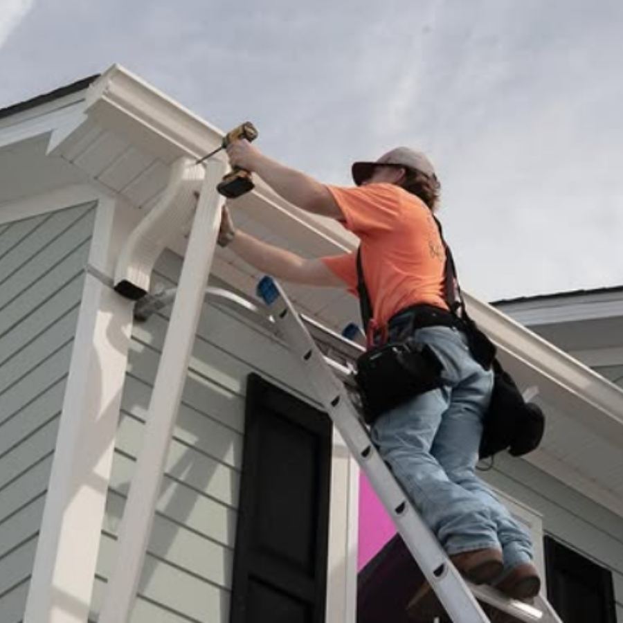 Gutter installation technician on ladder using drill to attach white seamless gutters and downspout on light gray siding home in Thunderbolt GA by Spartina Seamless Gutters.